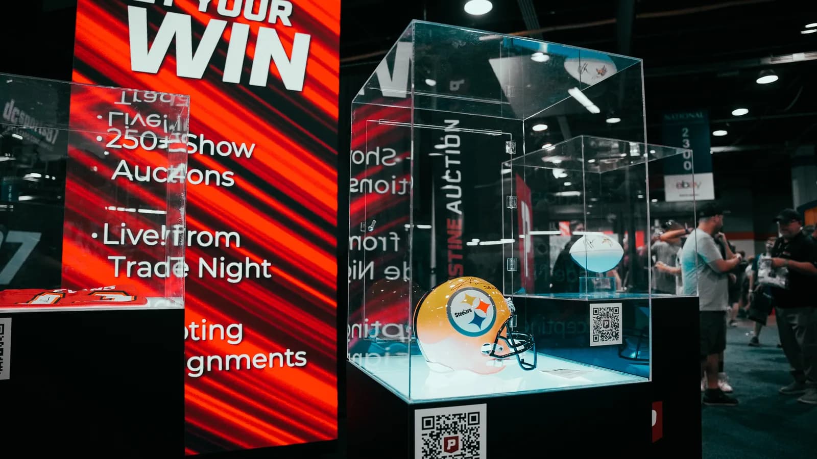 A signed Pittsburgh Steelers helmet lit inside an acrylic auction case at the Pristine Auction stand, beside a screen advertising 250+ show auctions live from Trade Night