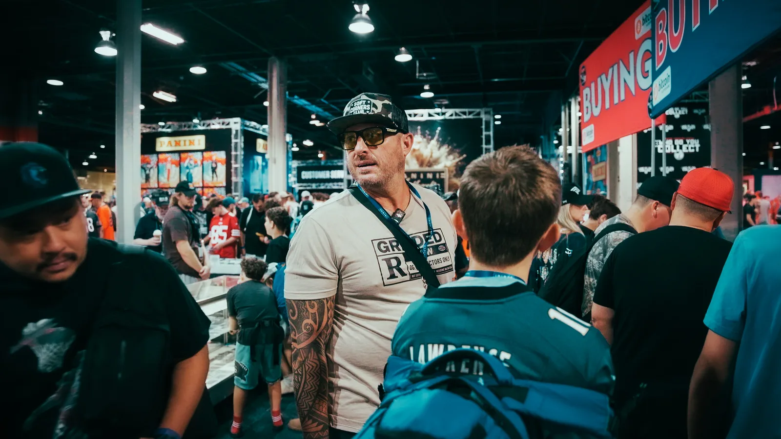 A crowded aisle at the 2026 National: a collector in a Graded shirt and camo cap mid-conversation, the illuminated Panini stand behind him, a dealer’s BUYING banner overhead, a child working a showcase to one side and a teenager in a Lawrence jersey passing in the foreground
